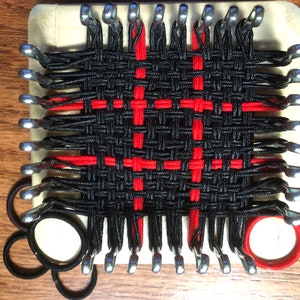 May include: A loom with black and red rubber bands woven into a pattern. The loom is made of wood and has metal pegs. There are extra rubber bands in the lower left corner of the image.