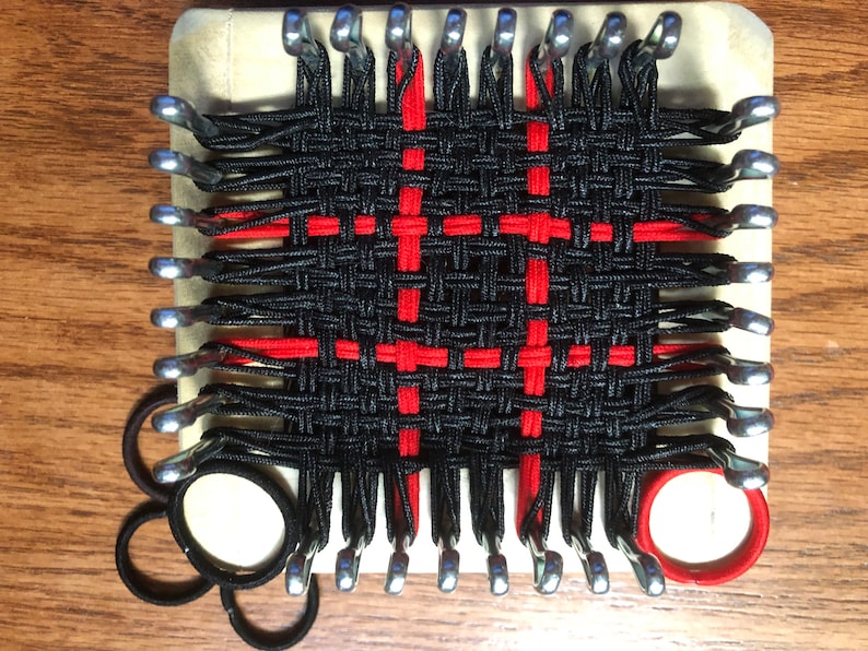 May include: A loom with black and red rubber bands woven into a pattern. The loom is made of wood and has metal pegs. There are extra rubber bands in the lower left corner of the image.