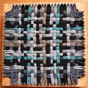 May include: A woven tapestry with a geometric pattern in shades of gray, black, brown, and teal. The tapestry is woven on a wooden loom with metal pins.