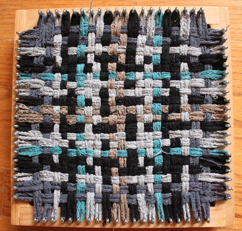 May include: A woven tapestry with a geometric pattern in shades of gray, black, brown, and teal. The tapestry is woven on a wooden loom with metal pins.