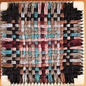 May include: A woven tapestry with a geometric pattern in shades of brown, black, teal, pink, and red. The tapestry is woven on a loom with wooden pegs.