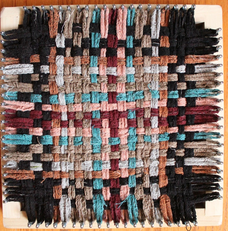 May include: A woven tapestry with a geometric pattern in shades of brown, black, teal, pink, and red. The tapestry is woven on a loom with wooden pegs.