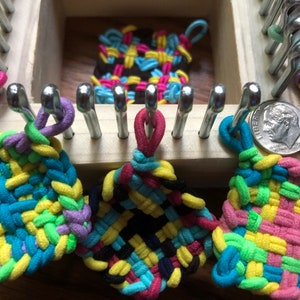 May include: Four colorful woven squares made with rubber bands on a wooden loom. The squares are in various colors, including pink, blue, green, yellow, and black.