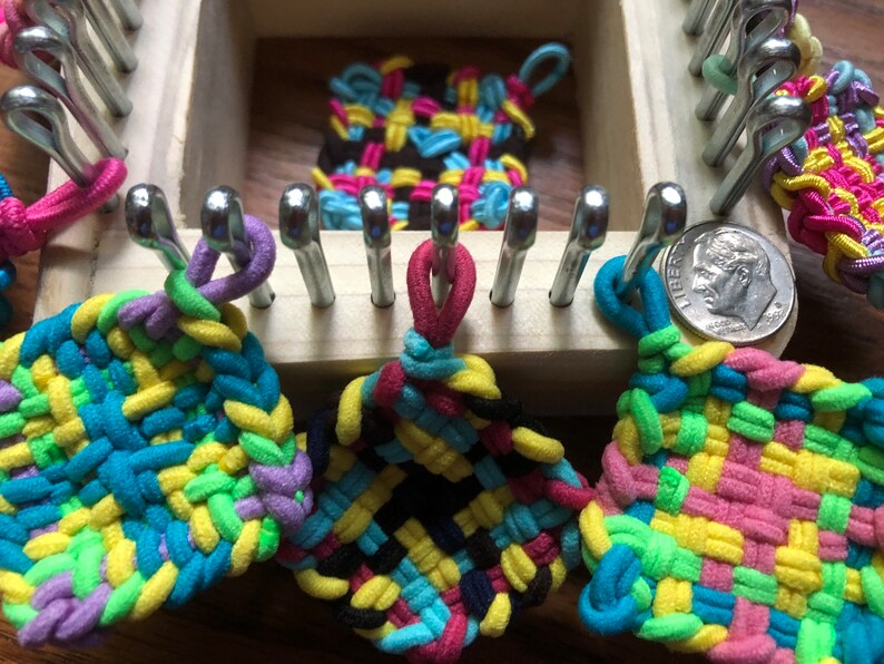 May include: Four colorful woven squares made with rubber bands on a wooden loom. The squares are in various colors, including pink, blue, green, yellow, and black.