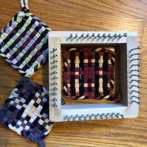 May include: Three woven pot holders, two finished and one in progress on a wooden loom. The finished pot holders are black, white, red, and beige. The pot holder on the loom is black, red, beige, and cream.