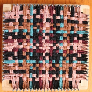 May include: A woven tapestry with a geometric pattern in black, brown, pink, and teal colors. The tapestry is woven on a loom with wooden frame and metal pegs.