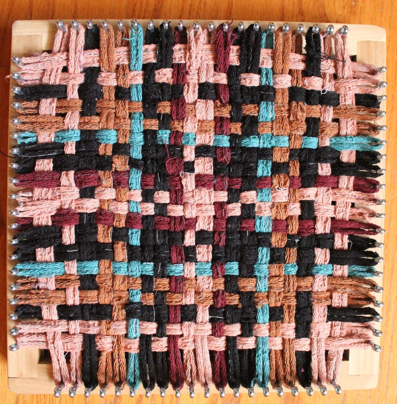 May include: A woven tapestry with a geometric pattern in black, brown, pink, and teal colors. The tapestry is woven on a loom with wooden frame and metal pegs.