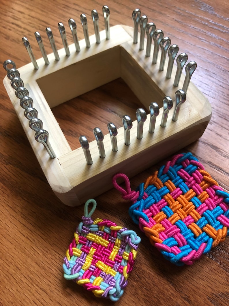 May include: A wooden loom with metal pegs for weaving colorful rubber band bracelets. Two finished bracelets are shown, one with pink, yellow, and purple bands, and the other with blue, orange, and pink bands.