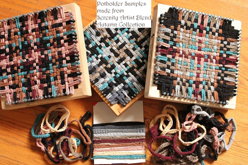 May include: Three wooden looms with woven potholders made from Serenity Artist Blend yarn in the Autumn Collection. The yarn is in various colors, including black, brown, teal, and pink. There are also loose strands of yarn in the same colors.