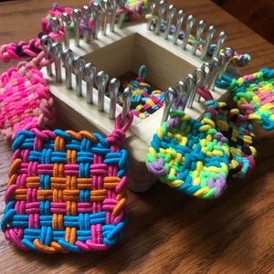 May include: A wooden loom with colorful rubber band bracelets and squares hanging from it. The loom has metal hooks and is used for making crafts.