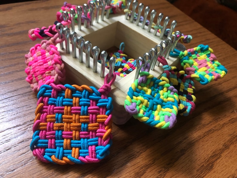May include: A wooden loom with colorful rubber band bracelets and squares hanging from it. The loom has metal hooks and is used for making crafts.