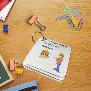 May include: A stack of white cards on a metal ring with the text "It is important to listen to my teachers." and cartoon illustrations. The cards are on a wooden surface with colorful paper clips and binder clips.