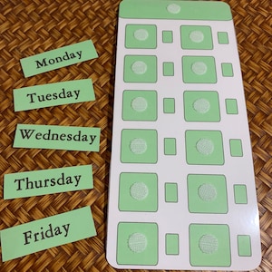 May include: A light green and white weekly planner with the days of the week printed on individual rectangular labels. The planner has square sections with a circular velcro patch in the center. The labels read Monday through Friday.