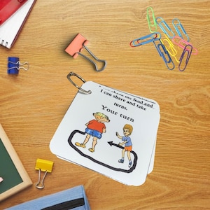 May include: A stack of white cards with a metal ring, featuring illustrations of children and text that reads "Your turn." The cards are on a wooden surface, surrounded by colorful paper clips and binder clips.