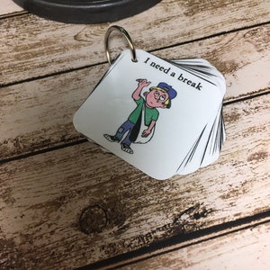 May include: A stack of white square cards held together by a metal ring. The top card has an illustration of a cartoon character with the text "I need a break". The character is wearing a green shirt, blue jeans, and a blue hat.