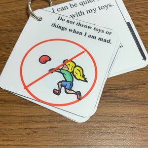 May include: A set of white cards with a silver ring, featuring text and illustrations. One card shows a child throwing a ball with a red circle and a line through it. Text reads, "I can be quiet and with my toys. Do not throw toys or things when I am mad."