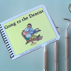 Può includere: Un libro a spirale intitolato "Going to the Dentist" con un'illustrazione di un dentista e di un paziente. Sono visibili due strumenti odontoiatrici. Il libro è su una superficie azzurra.