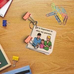 May include: A small notepad with a family illustration and the text "It is important to obey my parents." is held together by two pink binder clips. Other office supplies, including paper clips and a blue binder clip, are scattered on a wooden surface.