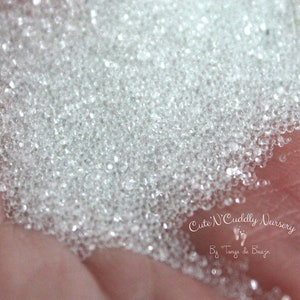 May include: A close-up of a hand holding a pile of clear glass beads. The beads are small and round, and they have a slight sparkle to them.