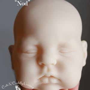 May include: A pale, unpainted baby doll head with closed eyes, labelled "Nod." The doll head is held by a hand and features a detailed face with a small mouth and defined features. The logo "Cute 'N Cuddly Nursery" is visible.