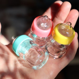 K&ouml;nnte beinhalten: Drei Miniatur-Glas-Babyflaschen mit rosa, gelben und t&uuml;rkisfarbenen Deckeln. Die Flaschen werden in einer Hand gehalten, wodurch ihre klare Glaskonstruktion und geringe Gr&ouml;&szlig;e hervorgehoben werden. Die Flaschen sind mit einer klaren Fl&uuml;ssigkeit gef&uuml;llt.