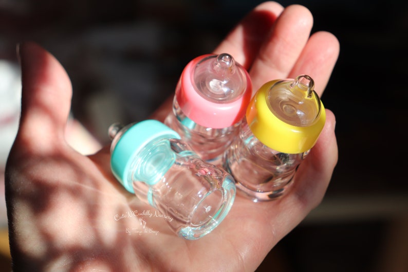 K&ouml;nnte beinhalten: Drei Miniatur-Glas-Babyflaschen mit rosa, gelben und t&uuml;rkisfarbenen Deckeln. Die Flaschen werden in einer Hand gehalten, wodurch ihre klare Glaskonstruktion und geringe Gr&ouml;&szlig;e hervorgehoben werden. Die Flaschen sind mit einer klaren Fl&uuml;ssigkeit gef&uuml;llt.