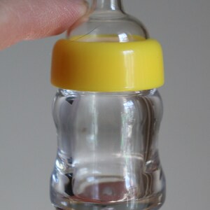 K&ouml;nnte beinhalten: Eine kleine, transparente Babyflasche mit gelbem Deckel. Die Flasche wird von den Fingern einer Person gehalten. Der Text "Cute 'N Cuddly Nursery" und "By Tanya de Bruijn" sind sichtbar.