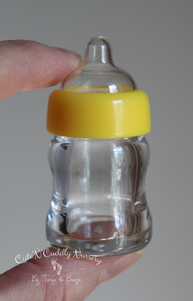 K&ouml;nnte beinhalten: Eine kleine, transparente Babyflasche mit gelbem Deckel. Die Flasche wird von den Fingern einer Person gehalten. Der Text "Cute 'N Cuddly Nursery" und "By Tanya de Bruijn" sind sichtbar.