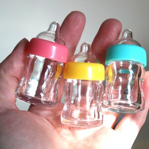 K&ouml;nnte beinhalten: Drei transparente Glas-Babyflaschen mit farbigen Deckeln in Rosa, Gelb und T&uuml;rkis. Die Flaschen werden in einer Hand gehalten, was ihre geringe Gr&ouml;&szlig;e und ihr Design hervorhebt. Geeignet f&uuml;r Babypartys.