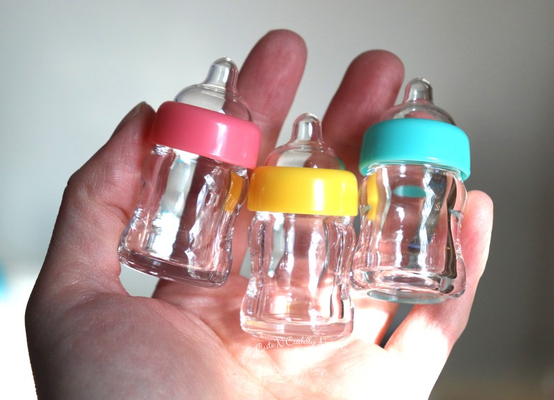 K&ouml;nnte beinhalten: Drei transparente Glas-Babyflaschen mit farbigen Deckeln in Rosa, Gelb und T&uuml;rkis. Die Flaschen werden in einer Hand gehalten, was ihre geringe Gr&ouml;&szlig;e und ihr Design hervorhebt. Geeignet f&uuml;r Babypartys.