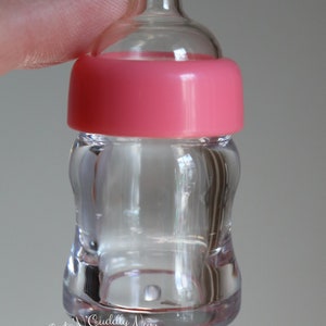 K&ouml;nnte beinhalten: Transparente Babyflasche mit rosa Kappe. Die Flasche hat eine geschwungene Form und einen Sauger oben. Der Text "Cute N' Cuddly Nursery" und "By Tanya de Bruijn" sind sichtbar.