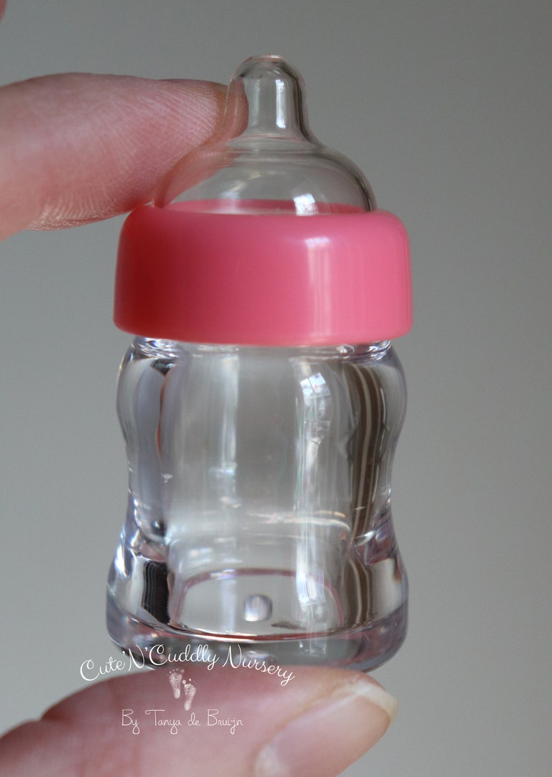 K&ouml;nnte beinhalten: Transparente Babyflasche mit rosa Kappe. Die Flasche hat eine geschwungene Form und einen Sauger oben. Der Text "Cute N' Cuddly Nursery" und "By Tanya de Bruijn" sind sichtbar.