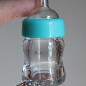K&ouml;nnte beinhalten: Eine transparente Babyflasche aus Glas mit einem t&uuml;rkisfarbenen Ring um den Hals. Die Flasche hat eine geschwungene Form und einen transparenten Sauger. Die Flasche wird von den Fingern einer Person gehalten. Der Text "Cute 'N Cuddly Nursery" ist sichtbar.