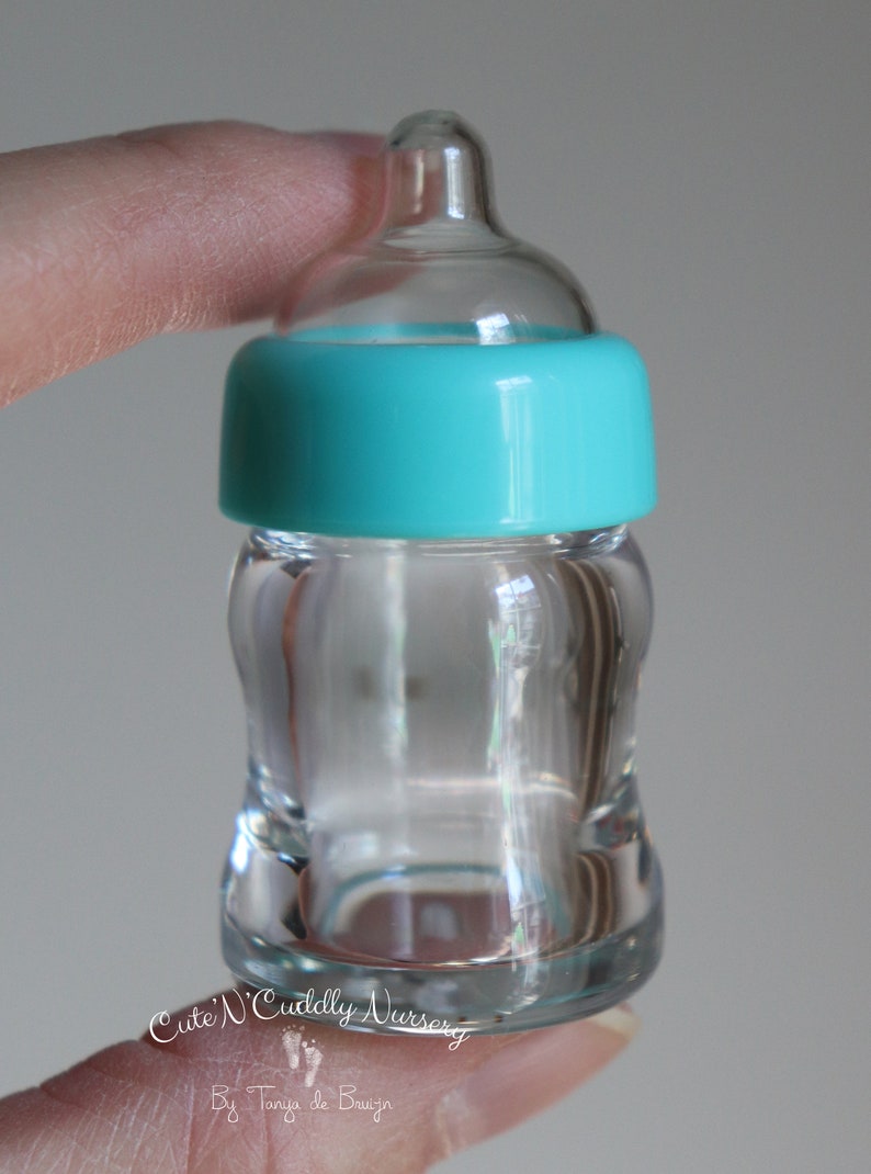 K&ouml;nnte beinhalten: Eine transparente Babyflasche aus Glas mit einem t&uuml;rkisfarbenen Ring um den Hals. Die Flasche hat eine geschwungene Form und einen transparenten Sauger. Die Flasche wird von den Fingern einer Person gehalten. Der Text "Cute 'N Cuddly Nursery" ist sichtbar.