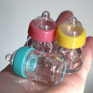K&ouml;nnte beinhalten: Drei Miniatur-Glas-Babyflaschen mit rosa, gelben und t&uuml;rkisfarbenen Deckeln. Die transparenten Flaschen haben farbige Deckel. Die kleinen Flaschen sind wahrscheinlich Dekorationsartikel.