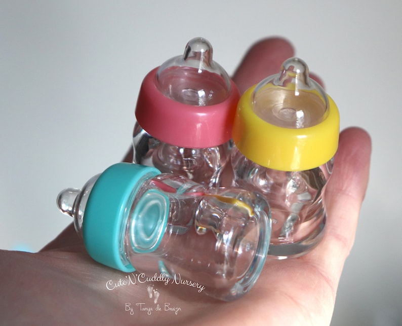 K&ouml;nnte beinhalten: Drei Miniatur-Glas-Babyflaschen mit rosa, gelben und t&uuml;rkisfarbenen Deckeln. Die transparenten Flaschen haben farbige Deckel. Die kleinen Flaschen sind wahrscheinlich Dekorationsartikel.