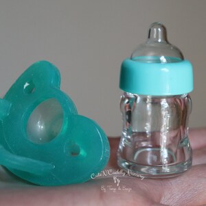 K&ouml;nnte beinhalten: Ein t&uuml;rkisfarbener Schnuller und eine transparente Babyflasche aus Glas mit t&uuml;rkisfarbenem Deckel. Der Schnuller befindet sich links und die Flasche rechts. Der Text "Cute 'N Cuddly Nursery" ist sichtbar.