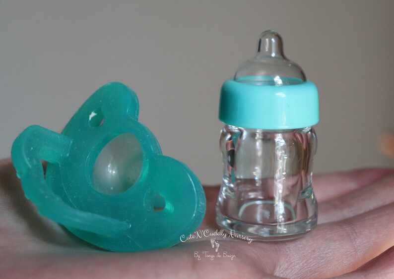K&ouml;nnte beinhalten: Ein t&uuml;rkisfarbener Schnuller und eine transparente Babyflasche aus Glas mit t&uuml;rkisfarbenem Deckel. Der Schnuller befindet sich links und die Flasche rechts. Der Text "Cute 'N Cuddly Nursery" ist sichtbar.