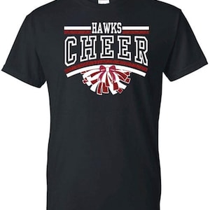 May include: A black short-sleeved t-shirt featuring a graphic print with the text HAWKS CHEER in white block letters. The design includes red glitter accents and a stylized illustration of a cheerleading pom-pom with a bow at the center.