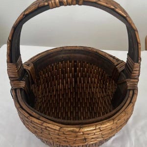 May include: A round, dark brown woven basket featuring a sturdy, arched handle attached to the sides. The basket is constructed with a tight, vertical weave pattern and a smooth, solid rim, showcasing a natural, polished wood-like finish throughout.