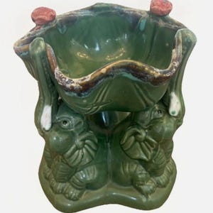 May include: A green ceramic bowl supported by three elephant figures. The bowl features a wavy, dark-rimmed edge with two small, reddish-brown decorative accents on the top rim. The elephants are sculpted in a seated position with trunks raised.