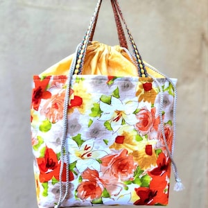 May include: A white drawstring bag with a floral print featuring orange, red, yellow, and green flowers. The bag has two long straps and a yellow lining.