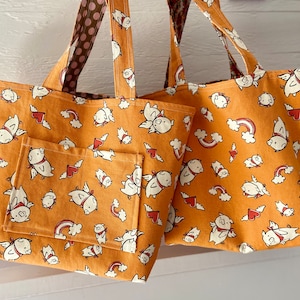 May include: Two brown canvas tote bags with a pink and white patterned fabric handle. Both bags feature a white and pink pig design with hearts and rainbows. The smaller bag has a pocket on the front.