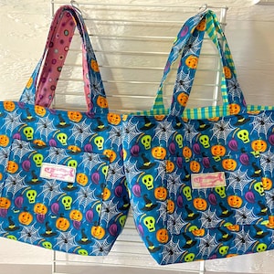 Może przedstawiać: Dwie niebieskie torby na zakupy na Halloween z wzorem czaszki, dyni i pajęczyny. Torby mają kieszeń z przodu z różową etykietą z napisem "CF-Designs".