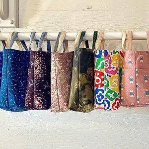 Puede incluir: Una colección de 14 bolsas de tela coloridas con varios patrones y diseños. Las bolsas están colgadas en un estante y están hechas de tela.