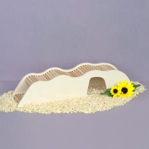 Könnte beinhalten: Eine helle Holzbrücke mit gewellter Oberkante und einer mittigen Bogenöffnung, die ein Design mit Lattensteg aufweist. Die Struktur steht auf einem Bett aus hellen Holzspänen und ist mit zwei leuchtend gelben Sonnenblumen verziert.