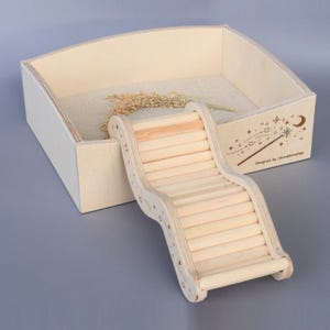Könnte beinhalten: Ein hölzernes Hamstergehege mit Rampe und sandgefüllter Basis. Das Gehege besteht aus hellem Holz und verfügt über eine gebogene Rampe für einen einfachen Zugang. Das Design beinhaltet eine dekorative Illustration eines Zauberstabs, Sterne und einen Mond.