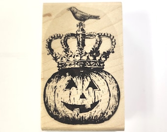 May include: A wooden stamp with a black and white illustration of a pumpkin wearing a crown with a bird perched on top. The pumpkin has a carved jack-o'-lantern face.
