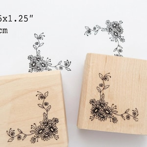 May include: Wooden stamp with a floral corner design. The stamp is 3x3cm and features a detailed illustration of daisies and leaves. The stamp is shown with its impression on white paper.
