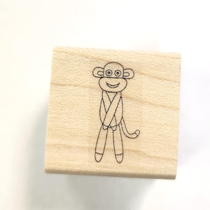 Pode incluir: Um carimbo de borracha de madeira com um desenho de linha preta de um macaco com olhos grandes e um sorriso. O macaco está vestindo uma camisa e tem uma cauda.